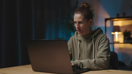 Focused woman in casual hoodie and eyewear sitting at desk and typing on modern laptop. Program development and remote work concept.
