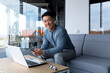 © Liubomir - Happy and successful asian working in office with laptop, businessman looking at camera and smiling, holding phone