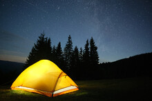 Bright Illuminated Tourist Tent Glowing On Camping Site In Dark Mountains Under Night Sky With Sparkling Stars. Active Lifestyle Concept