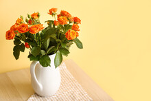 Jug With Beautiful Orange Roses On Table Against Yellow Wall