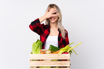 Farmer girl holding a basket full of fresh vegetables over isolated white background covering eyes by hands and smiling