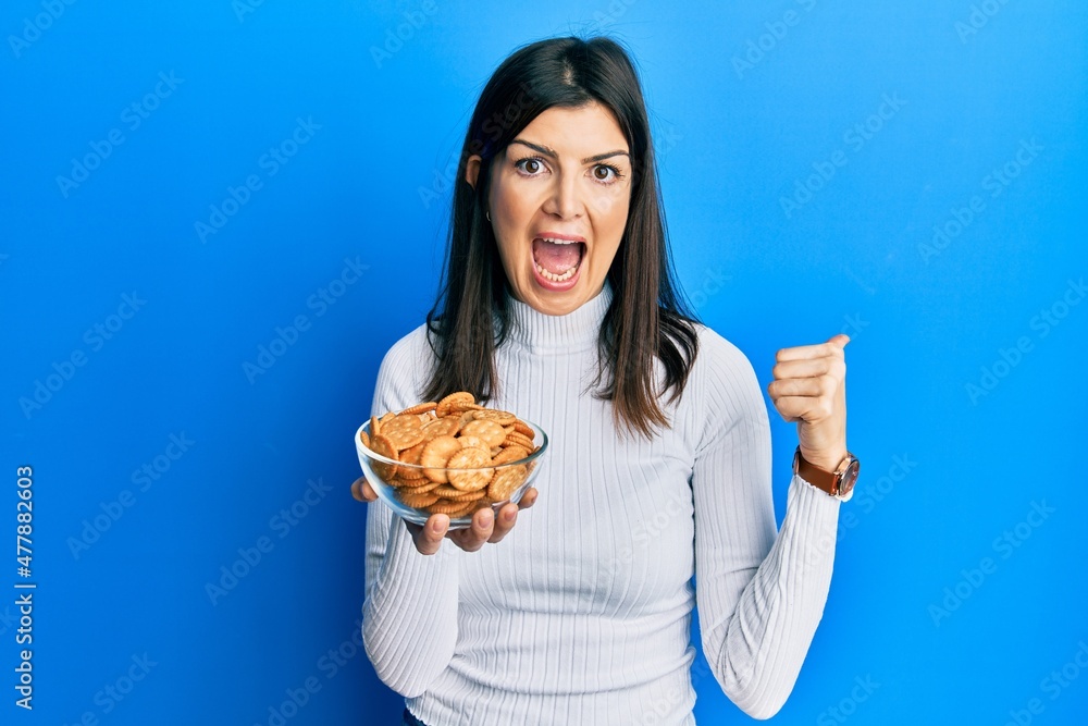 Young hispanic woman holding salty biscuits bowl pointing thumb up to the side smiling happy with open mouth