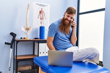 Canvas Print - Young redhead man wearing physiotherapist uniform using laptop and talking on the smartphone at physiotherapy clinic