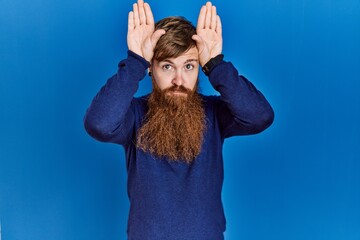 Poster - Redhead man with long beard wearing casual blue sweater over blue background doing bunny ears gesture with hands palms looking cynical and skeptical. easter rabbit concept.