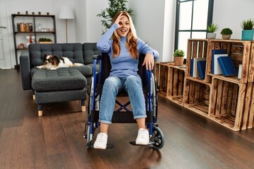 Poster - Young beautiful woman sitting on wheelchair at home doing ok gesture with hand smiling, eye looking through fingers with happy face.