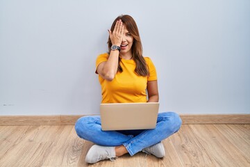 Poster - Hispanic woman using laptop sitting on the floor at home covering one eye with hand, confident smile on face and surprise emotion.