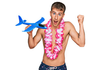 Wall Mural - Young blond man wearing swimsuit and hawaiian lei holding airplane toy screaming proud, celebrating victory and success very excited with raised arms