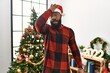 African american man wearing santa claus hat standing by christmas tree surprised with hand on head for mistake, remember error. forgot, bad memory concept.