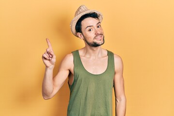 Canvas Print - Young hispanic man wearing summer hat with a big smile on face, pointing with hand and finger to the side looking at the camera.