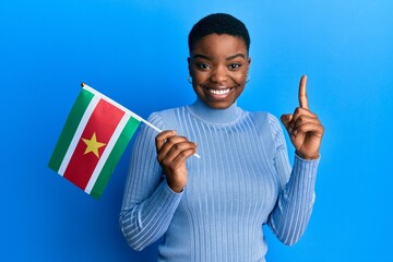 Sticker - Young african american woman holding suriname flag smiling with an idea or question pointing finger with happy face, number one