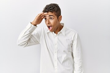 Wall Mural - Young handsome hispanic man standing over isolated background very happy and smiling looking far away with hand over head. searching concept.