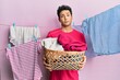© Krakenimages.com - Handsome hispanic man doing laundry holding wicker basket in shock face, looking skeptical and sarcastic, surprised with open mouth