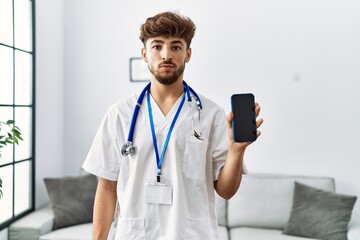 Canvas Print - Young arab man wearing doctor uniform and stethoscope holding smartphone thinking attitude and sober expression looking self confident