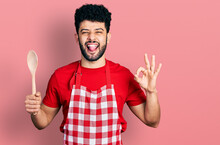 Young Arab Man With Beard Wearing Baker Uniform Holding Wooden Spoon Sticking Tongue Out Happy With Funny Expression.