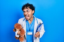 Young Hispanic Man Wearing Doctor Uniform Holding Teddy Bear And Clipboard Angry And Mad Screaming Frustrated And Furious, Shouting With Anger. Rage And Aggressive Concept.
