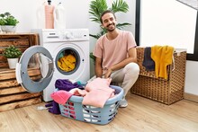 Young Hispanic Man Putting Dirty Laundry Into Washing Machine Looking Away To Side With Smile On Face, Natural Expression. Laughing Confident.