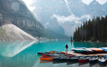 Tiny Person Standing On Dock With Colourful Canoes At Turquoise Blue Lake Surrounded By Mountains