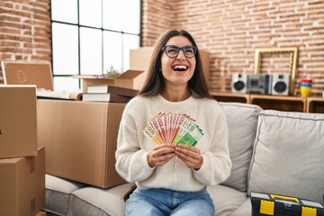 Young hispanic woman sitting on the sofa at new home holding money angry and mad screaming frustrated and furious, shouting with anger looking up.