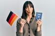 © Krakenimages.com - Young hispanic woman holding germany flag and passport making fish face with mouth and squinting eyes, crazy and comical.