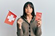 © Krakenimages.com - Young hispanic woman holding swiss flag and passport puffing cheeks with funny face. mouth inflated with air, catching air.