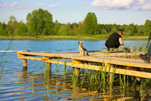 Woman With Small Yorkshire Terrier Dog Standing On Wooden Bridge, Pier, Footbridge Near River, Lake At Sunny Summer Day. Nature Recreation. Beautiful Landscape. Green Plants, Grass, Trees And Blue Sky
