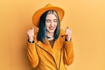 Young modern girl wearing yellow hat and leather jacket excited for success with arms raised and eyes closed celebrating victory smiling. winner concept.