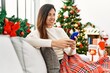 © Krakenimages.com - Middle age hispanic woman using smartphone sitting by christmas tree at home