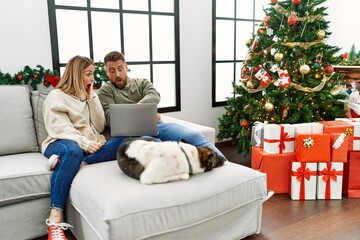 Sticker - Young couple using laptop sitting by christmas tree looking at the watch time worried, afraid of getting late