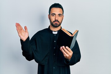 Poster - Handsome hispanic priest man with beard holding bible and christian cross relaxed with serious expression on face. simple and natural looking at the camera.