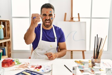 Sticker - Young hispanic man with beard at art studio with painted face angry and mad raising fist frustrated and furious while shouting with anger. rage and aggressive concept.