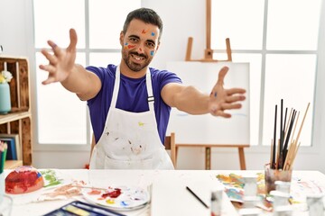 Sticker - Young hispanic man with beard at art studio with painted face looking at the camera smiling with open arms for hug. cheerful expression embracing happiness.