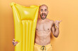 © Krakenimages.com - Young bald man wearing swimsuit and holding summer mattress float pointing thumb up to the side smiling happy with open mouth