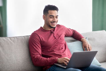 Wall Mural - Cheerful arab guy sitting on couch at home, using laptop