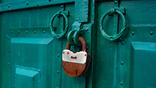 The Metal Gate Is Padlocked. Rusty Padlock On The Gate, Close-up. Rough Iron Doors.
