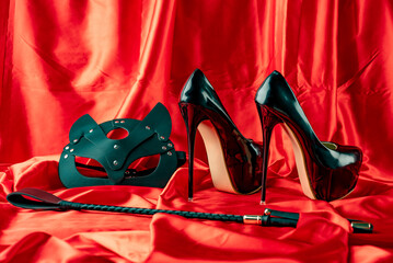 Adult sex games. BDSM items. Patent Fetish Extreme High Heel Stiletto Heels catr mask and whip on a red satin sheet.