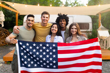 Diverse Millennial Friends Holding American Flag And Smiling At Camera In Front Of Motorhome At Campsite