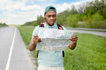 Unhappy Young Black Man Hitchhiking On Road, Looking At Map, Feeling Lost, Traveling Alone By Autostop
