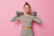 Horizontal Shot Of Satisfied Motivated Sportswoman Has Regular Training Raises Arms With Dumbbells Does Exercises For Muscles Dressed In Sportswear Smiles Toothily Isolated On Pink Wall. Sport Concept