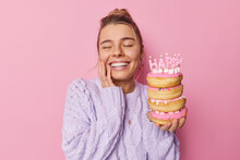 Optimistic European Woman Smiles Broadly Closes Eyes From Happiness Celebrates Special Occasion Holds Pile Of Asty Doughnuts Dressed In Knitted Jumper Isolated Over Pink Background. Happy Birthday