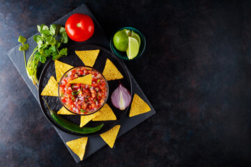 Wall Mural - Mexican Pico de Gallo salsa and ingredients. Bowl of tasty Pico de Gallo salsa and nachos on dark background. Top view. Copy space