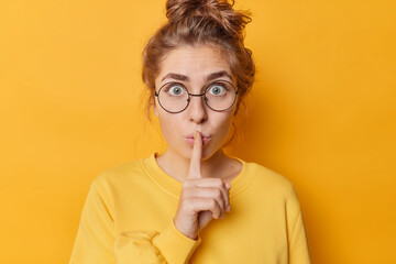 Stunned emotional beautiful woman hushing with index finger shares secret makes taboo gesture stares through round spectacles wears casual jumper isolated over yellow background. Shh be quiet