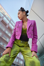 Vertical Shot Of Stylish Woman Dressed In Pink Jacket And Green Trouers Leans On Knees Focused Away Poses Against Urban Buildings In City Notices Something. People Fashion And Lifestyle Concept