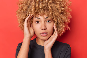 Poster - Close up portrait of self confident serious woman with curly blonde hair touches face looks directly at camera wears casual black t shirt isolated on red background shows natural beauty without makeup