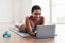 Smiling Young Black Woman Lying On Yoga Mat Using Laptop