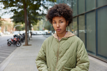 Wall Mural - Horizontal shot of serious attentive curly haired woman dressed in casual jacket strolls outdoors at city street stands against blurred urban background enjoys spare time. People and lifestyle concept