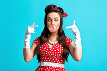 Beautiful Pinup Woman In Classic Outfit Blowing Bubble, Pointing At It On Blue Studio Background