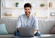 © Prostock-studio - Freelance job concept. Cheerful arab businessman working on laptop online and smiling sitting on sofa at home