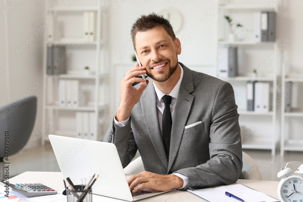 Smiling businessman with laptop talking by mobile phone at table in office