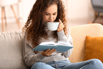 Sticker - Young African-American woman drinking tea while reading book on sofa at home