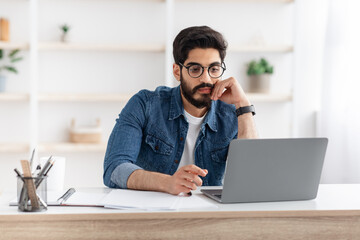 Young arab employee working on laptop at desk in home office, looking at computer screen, copy space